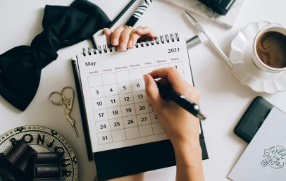 Top view of hands using a planner with coffee and accessories, focused on May 2021 calendar.